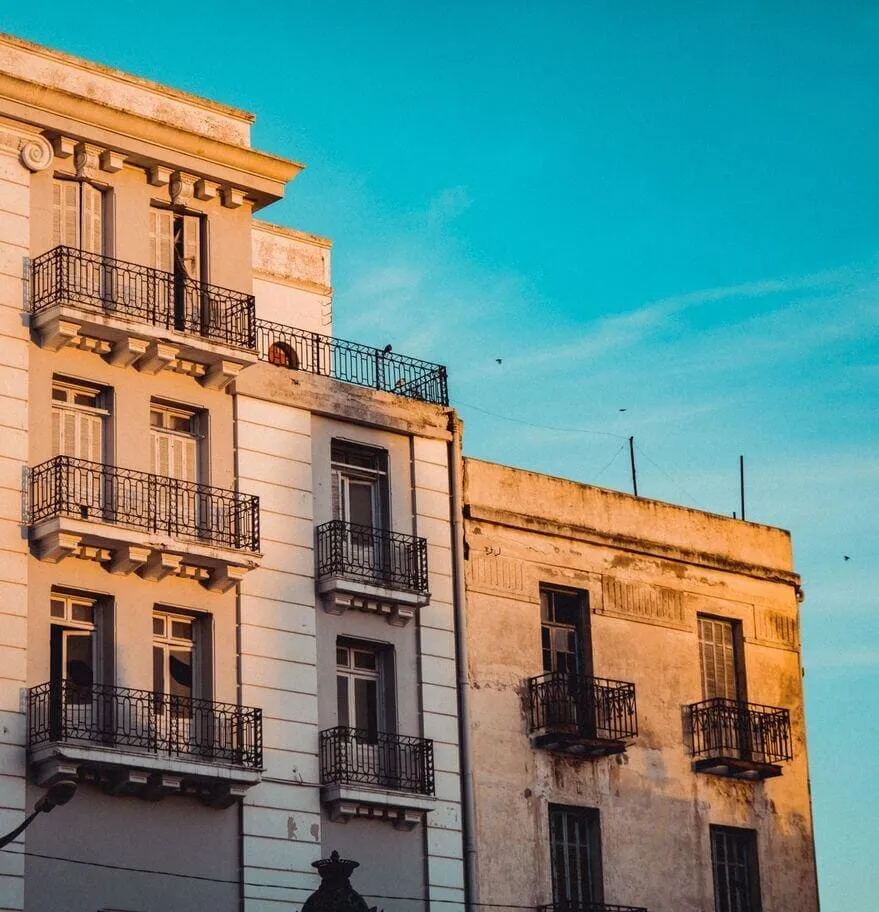 Two old buildings on a street in Tunis, wrought-iron balconies along their fronts, under a clear sky.
