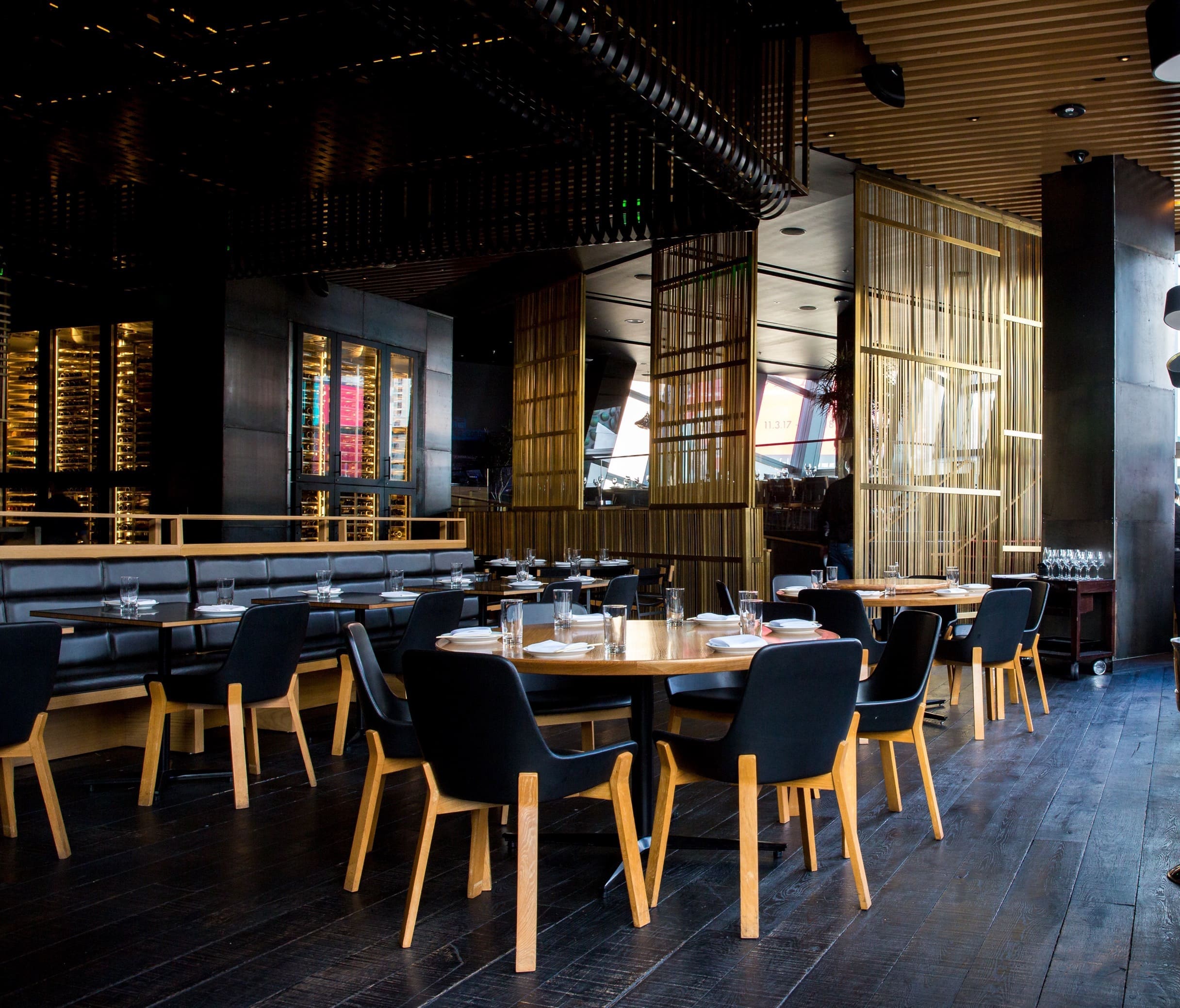 A restaurant dining room with black leather chairs, oak tables and brass-slat screens, its wine cellar lit behind glass.